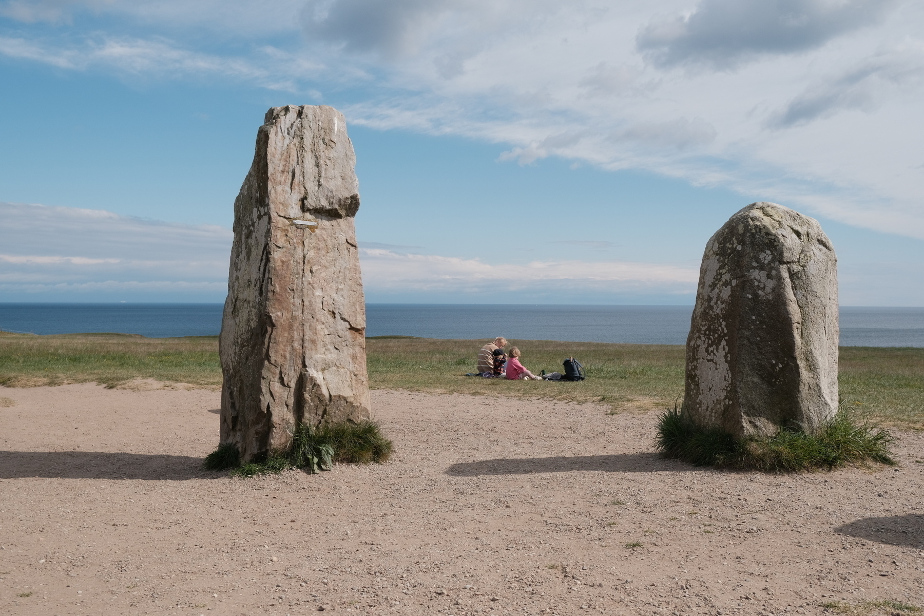 Två stora stenar står på en strand nära havet medan en familj sitter och vilar på marken i bakgrunden.